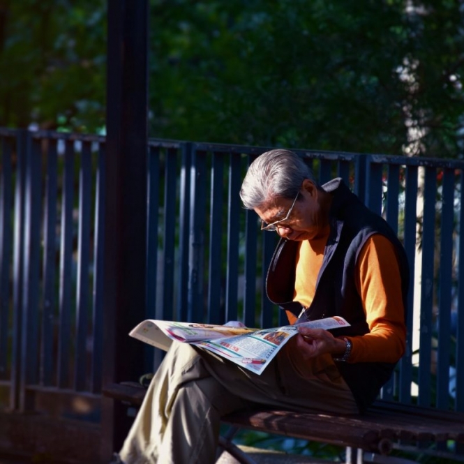 man reading newspaper while sitting on bench in front of wooden fence