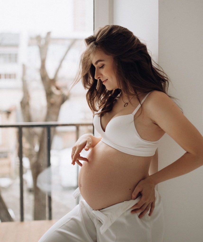 A pregnant woman in a white bra and trousers touching her belly by a window